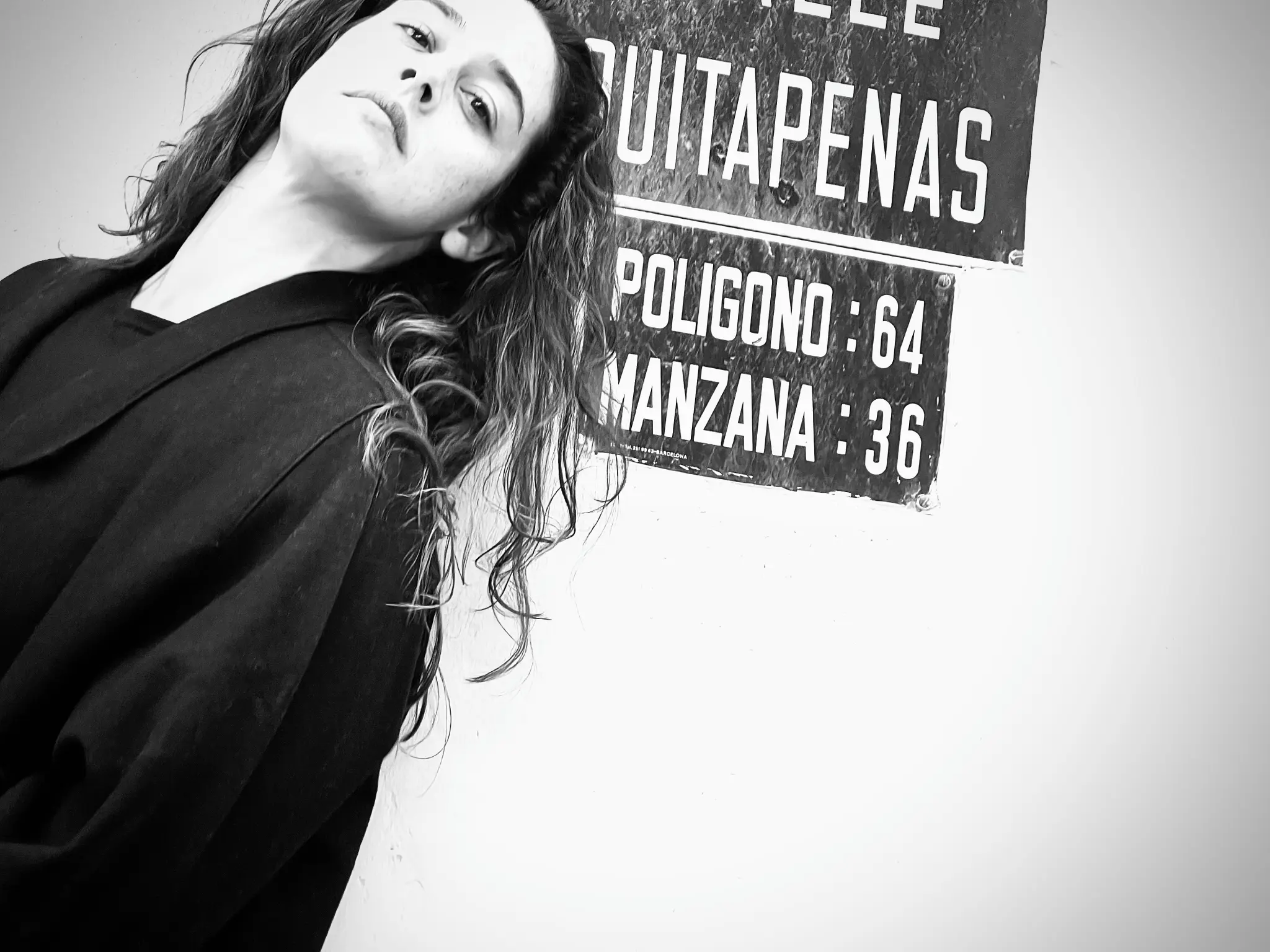 Alba Berrocal apoyada en una pared junto a un cartel esmaltado antiguo con la palabra Quitapenas, fotografía en blanco y negro