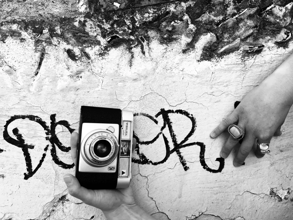 Dos manos sobre un muro agrietado, una sostiene una cámara de fotos antigua, fotografía en blanco y negro